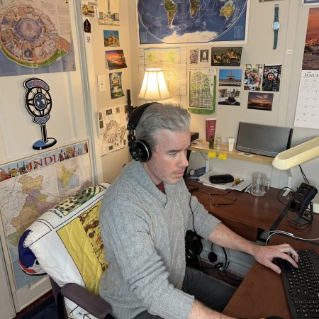 Podcast editor sitting at desk, with editing tools and maps on the wall.